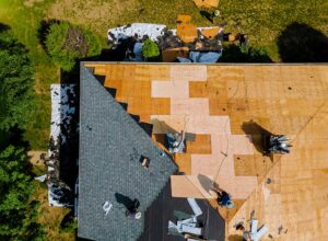 A person securing a blue tarp over damaged roof shingles during overcast weather.