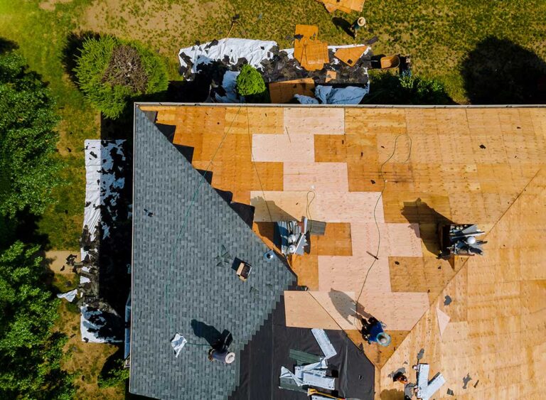 A person securing a blue tarp over damaged roof shingles during overcast weather.