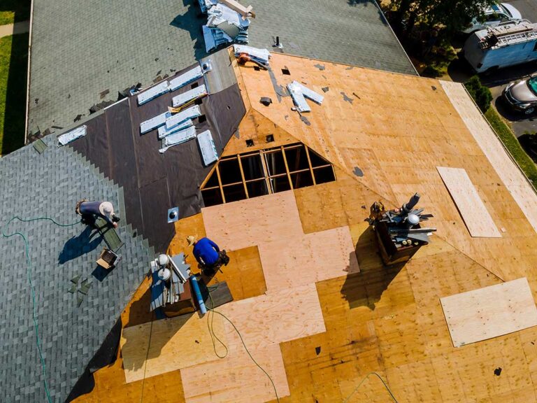A roofer examines labeled shingles, underlayment, and roof decking beside a residential chimney.